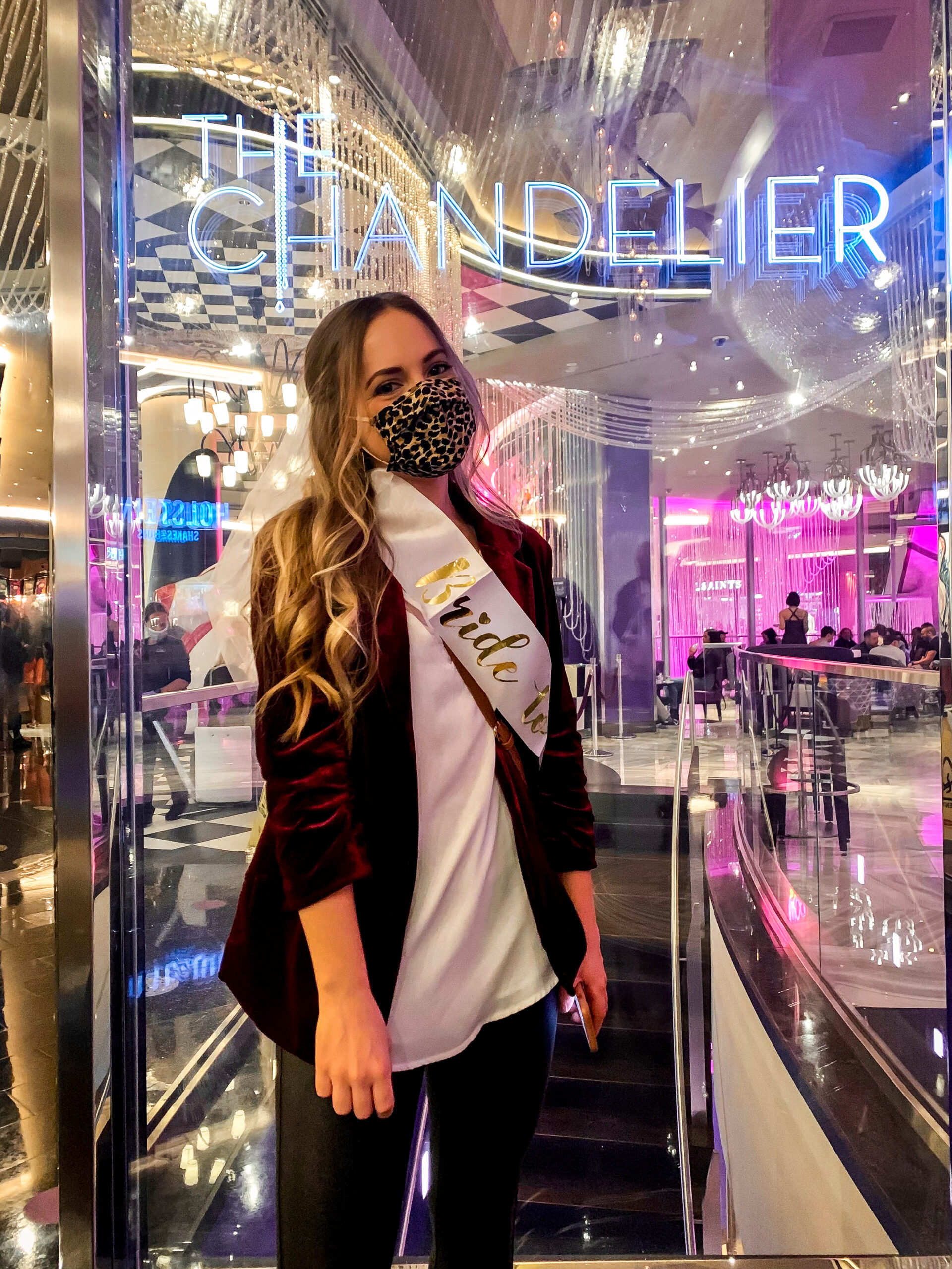 Blonde girl with mask standing in from of The Chandelier bar entry sign at the Cosmopolitan Hotel in Las Vegas for a bachelorette party.